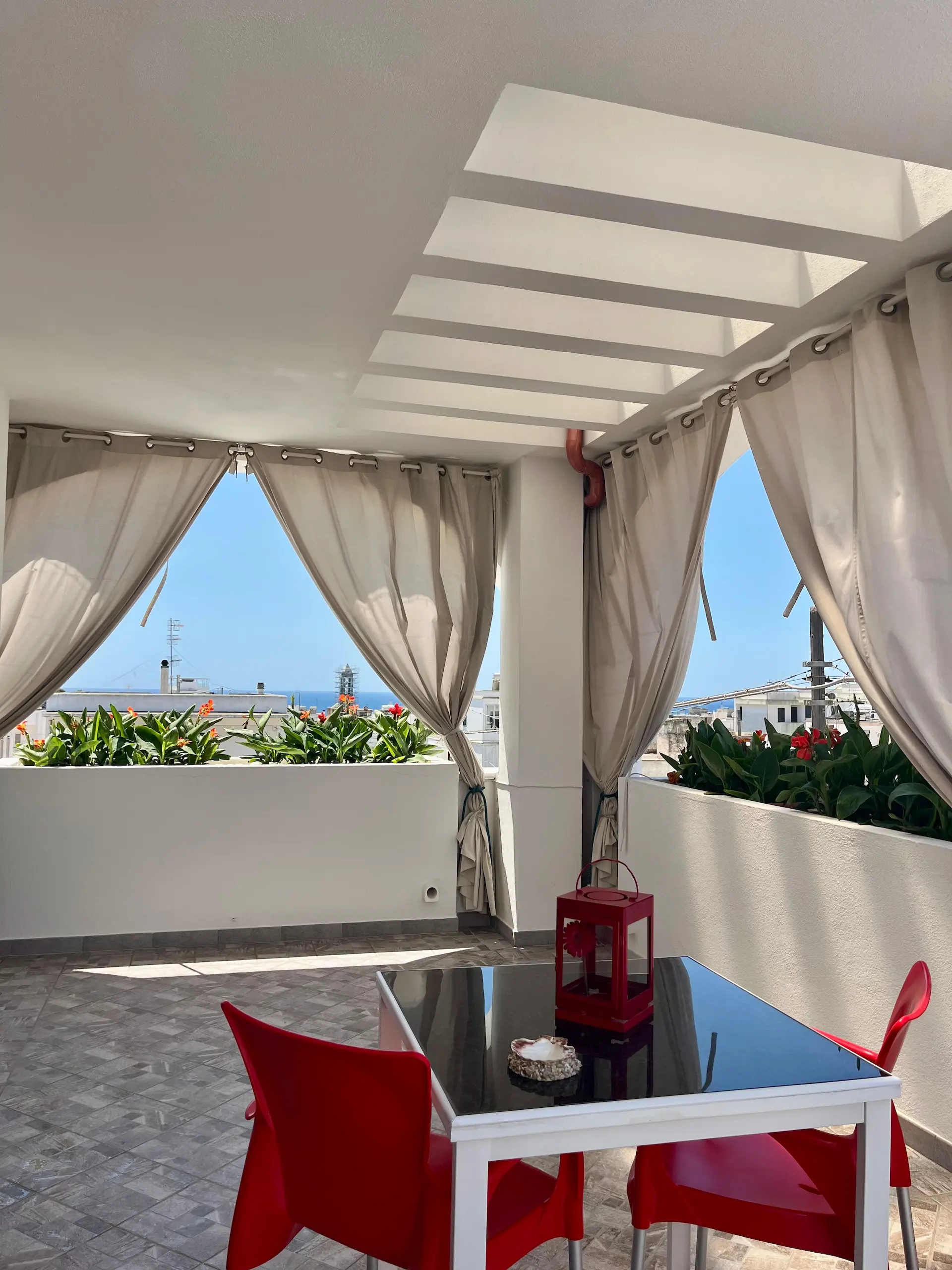 Veranda panoramica con fiori, tende beige, tavolo nero e sedie rosse con vista mare e vista su Leuca